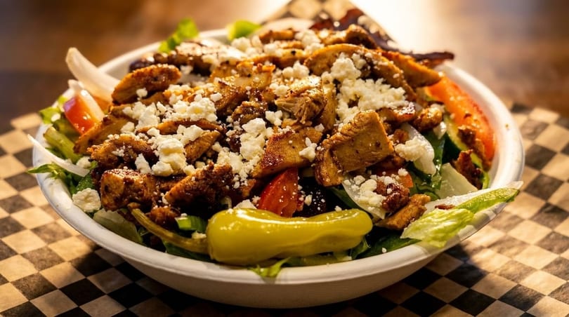 A close-up of a vibrant salad with grilled chicken, feta cheese, and a pepperoncini.