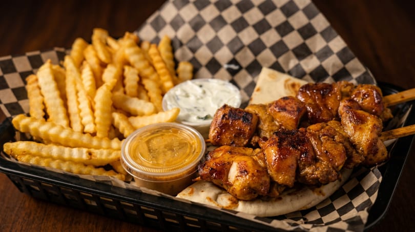 Chicken skewers with fries, pita bread, and two dipping sauces in a black basket.